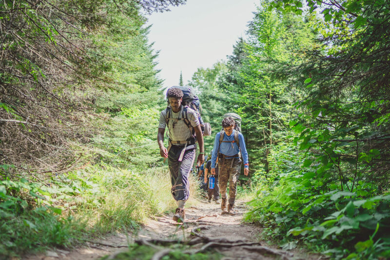 The image shows two hikers walking on a trail through a forest. They are both wearing backpacks and appear to be on a long hike. The trail is surrounded by trees and lush vegetation, suggesting a natural and scenic environment. The lighting indicates it is daytime.